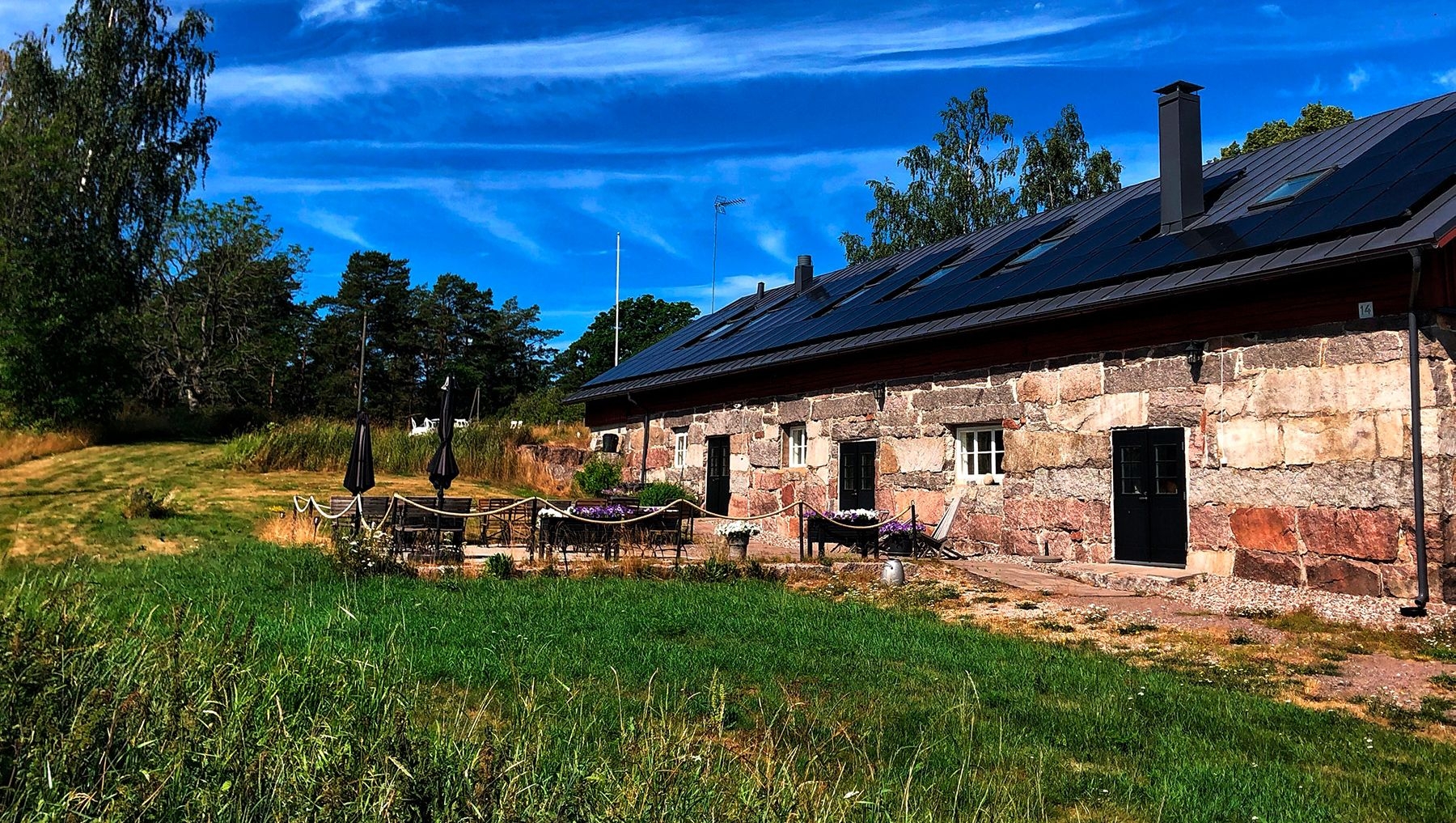 Bord och stolar på framsidan av Hotel Stallbacken, en stenbyggnad omgiven av gröna träd och en klarblå himmel.