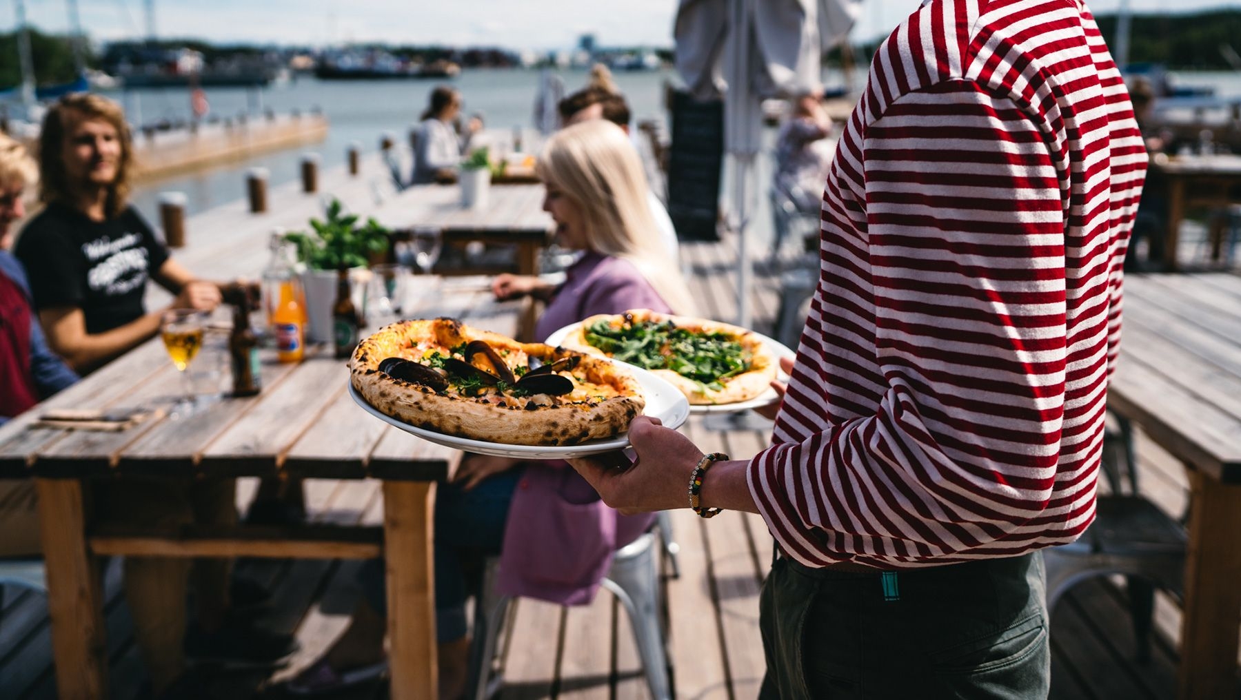 En servitör i en röd-vitrandig skjorta bär pizzor till matgäster på Zake Pizzeria & Wine Bar.