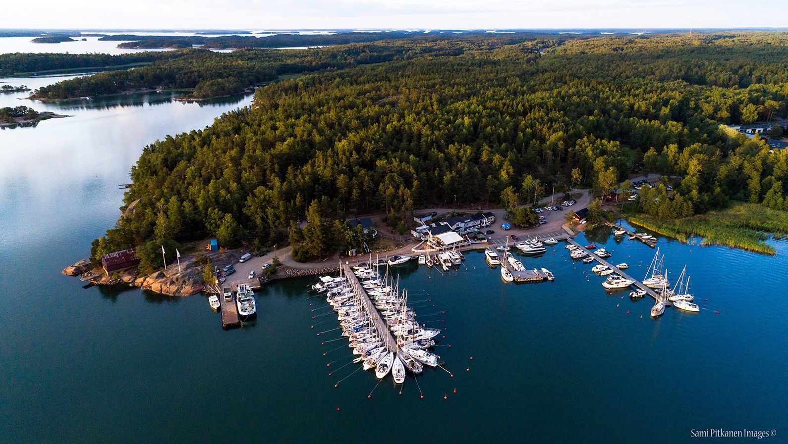 En flygbild över en båthamn. Många båtar har lagt till här, och det ligger bredvid en stor, grön skog.