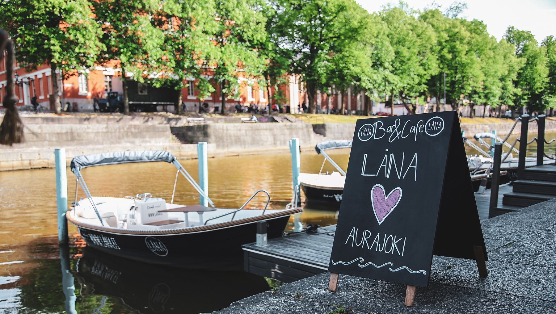 En svart tavla med reklam för Låna Bar & Café står vid Auraälven, där Lånabåtar har lagts till.