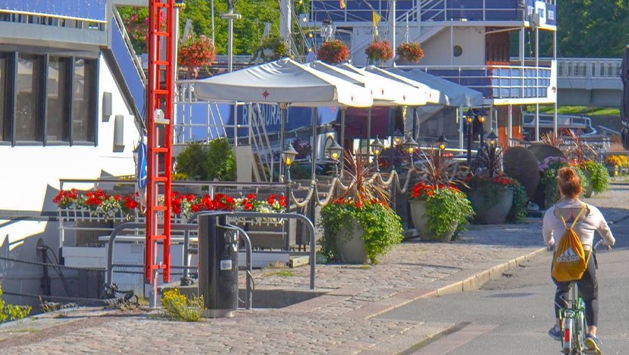 En cyklist cyklar på stigen bredvid Svarte Rudolf och andra flodbåtar, förankrade i Auraälven.