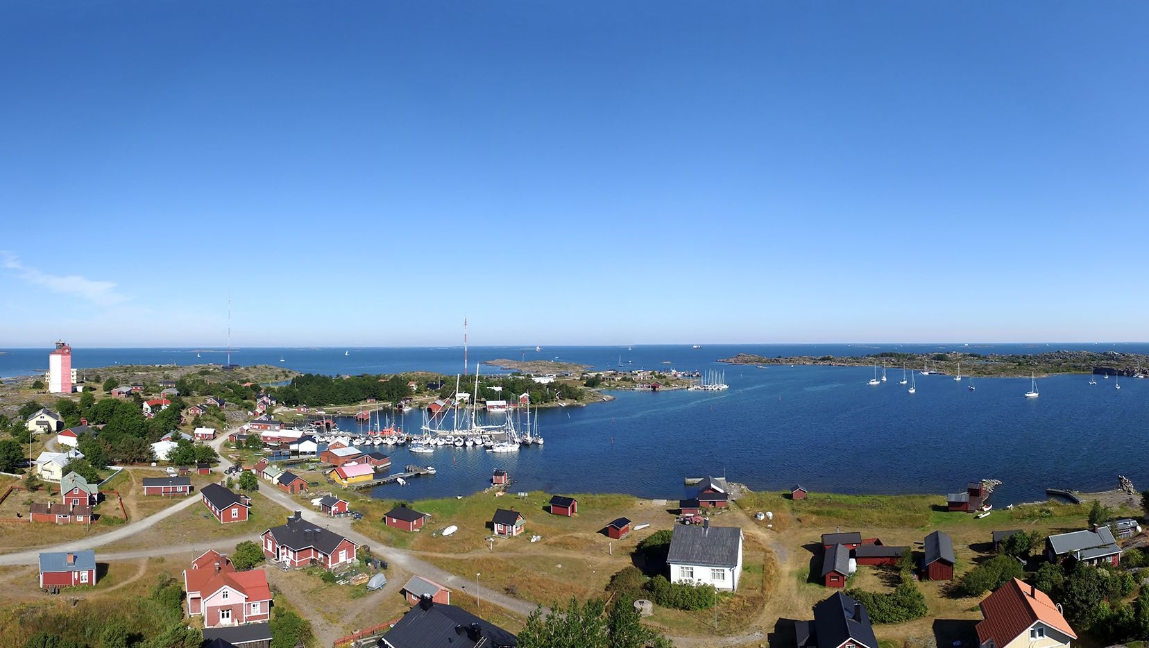 Ilmakuva Utön saarelta. Muutama purjevene on laiturissa ja kuvantaustalla on punavalkoinen majakka.