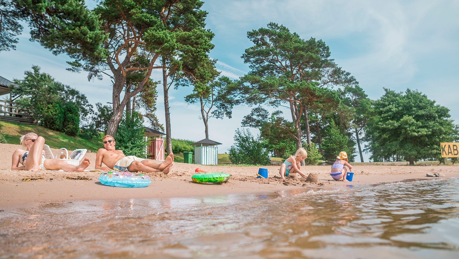 Två föräldrar kopplar av på sanden vid Kasnäs Beach, medan två barn bygger sandslott.