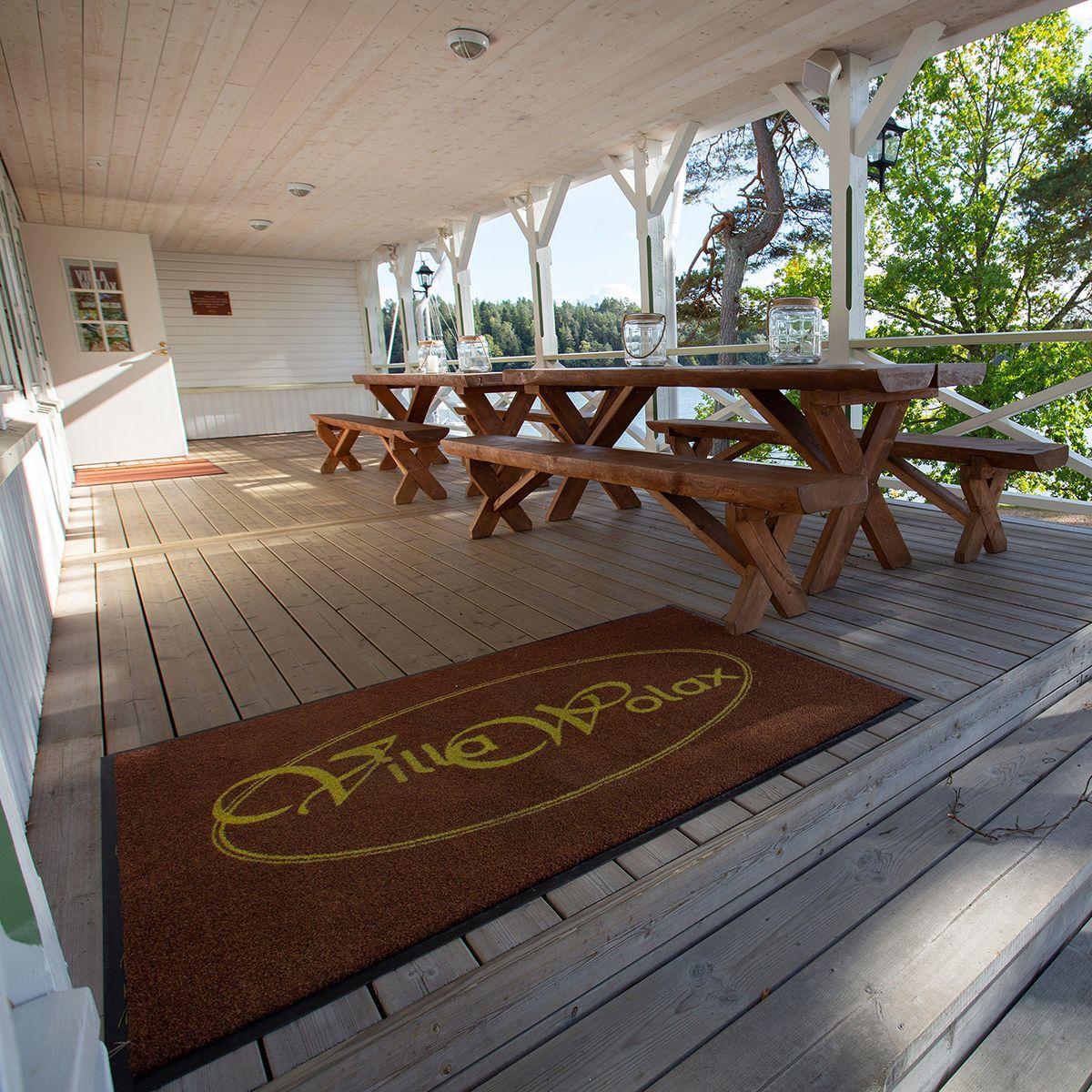 En matta med texten "Villa Wolax" ligger på golvet på en terrass, som också har ett långt träbord och havsutsikt.