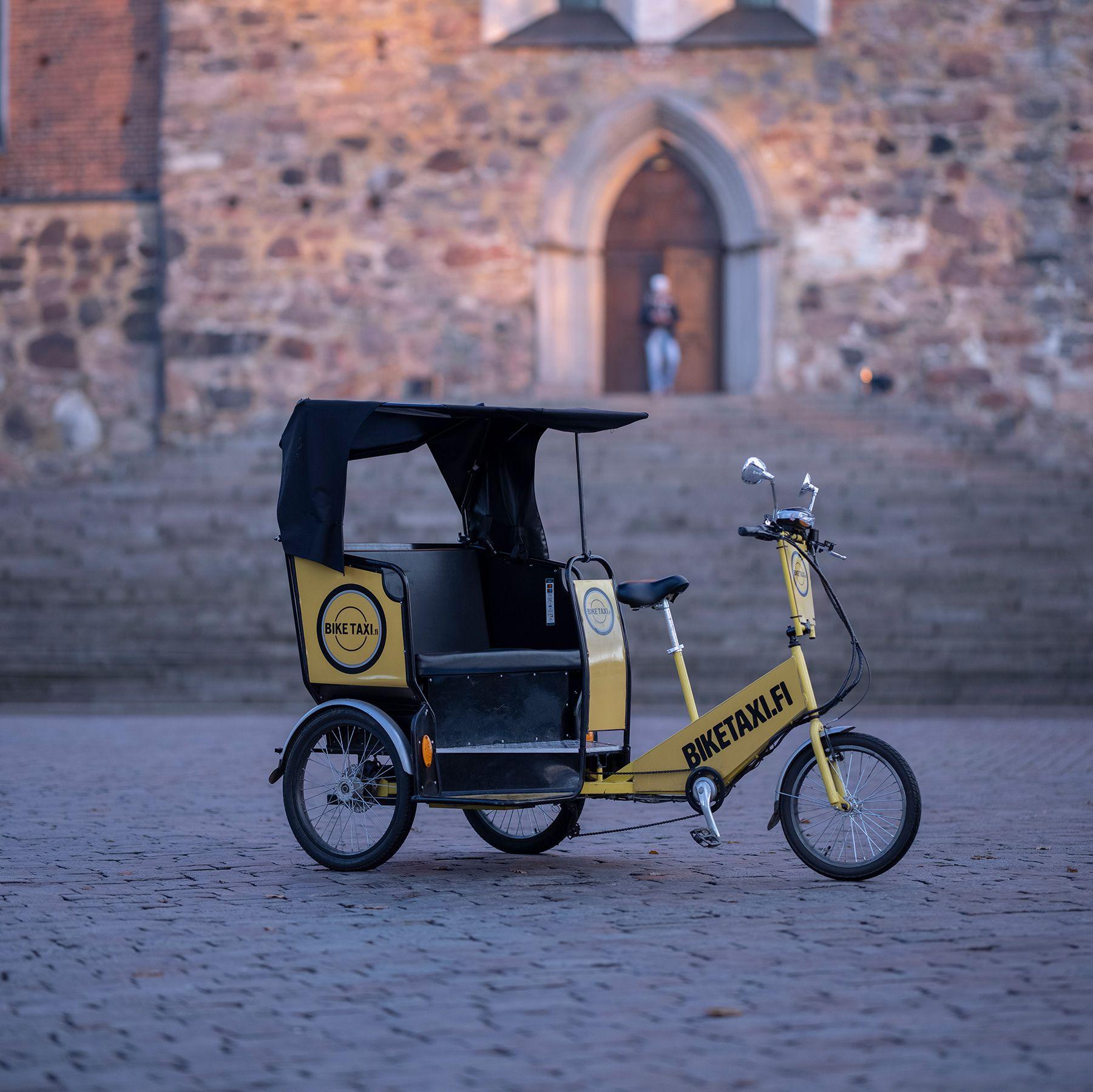 En elektrisk rickshaw som drivs av Biketaxi Åbo. Den är gul och svart och har tre hjul.