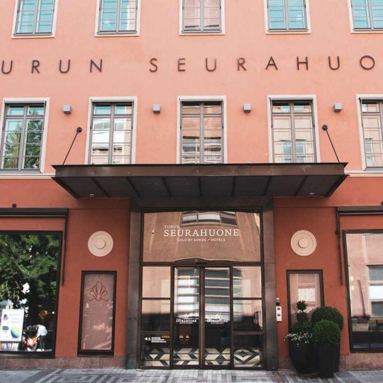 Den ljusrosa exteriören på Turun Seurahuone, med fyra flaggor och orden "Turun Seurahuone" på väggen.