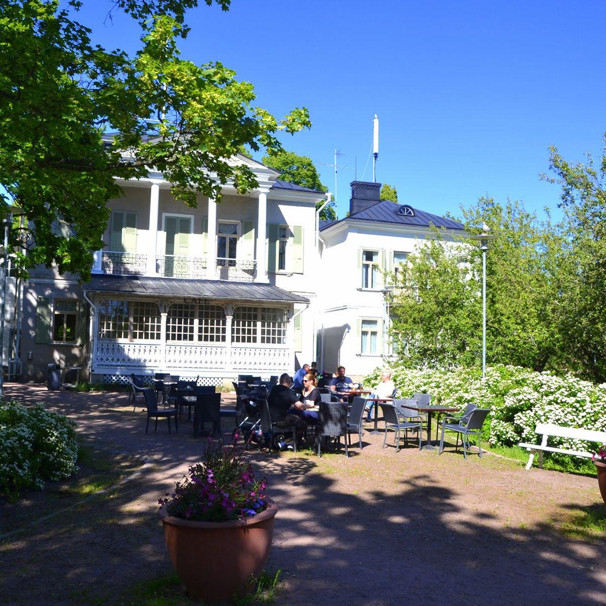 Vierailijat istuvat auringossa viehättävän vaalean, kaksikerroksisen huvilan edustalla Ruissalo Campingissa.