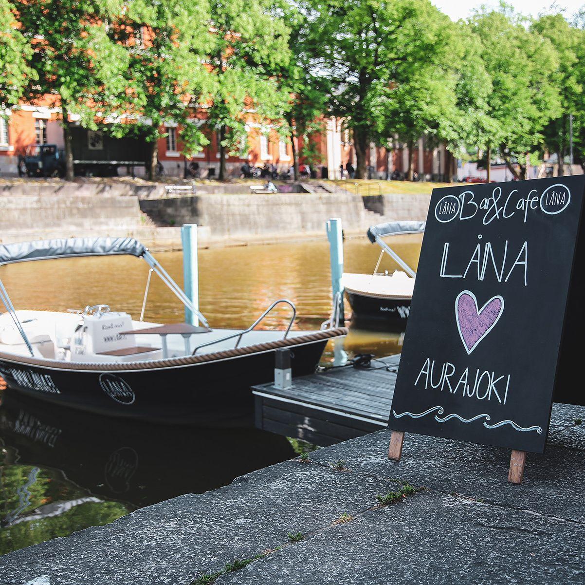 A chalkboard advertising Låna Bar & Café stands beside the Aura River, where Låna boats have been docked.
