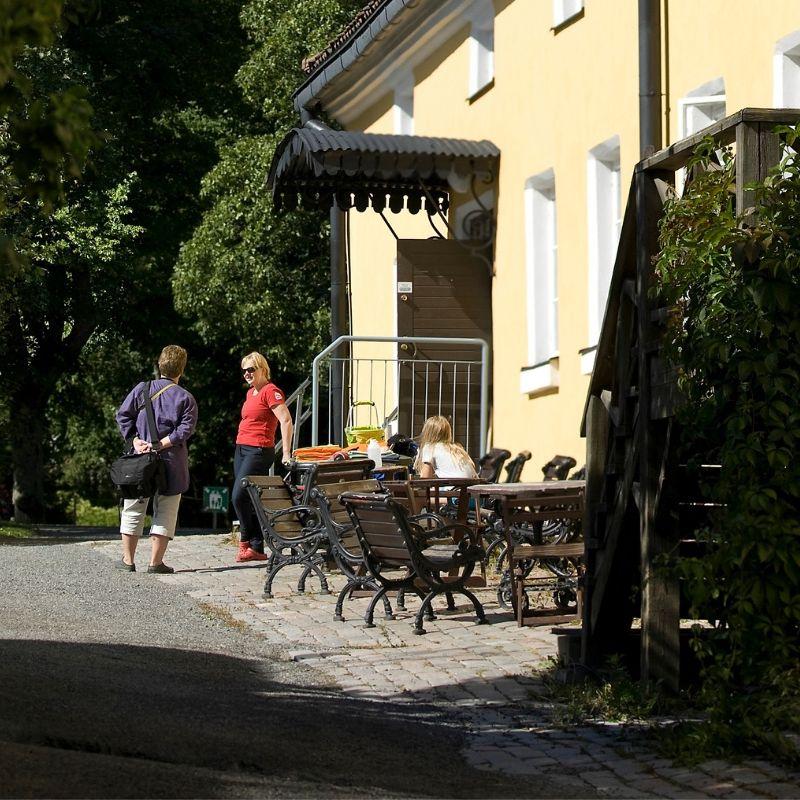 Two visitors stand outside the café at Hostel Tuorla, a yellow building with tables and chairs placed outside.