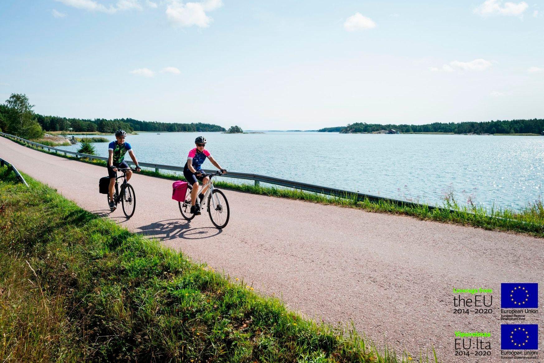 Två cyklister cyklar längs en landsväg. I bakgrunden syns den finska skärgården.
