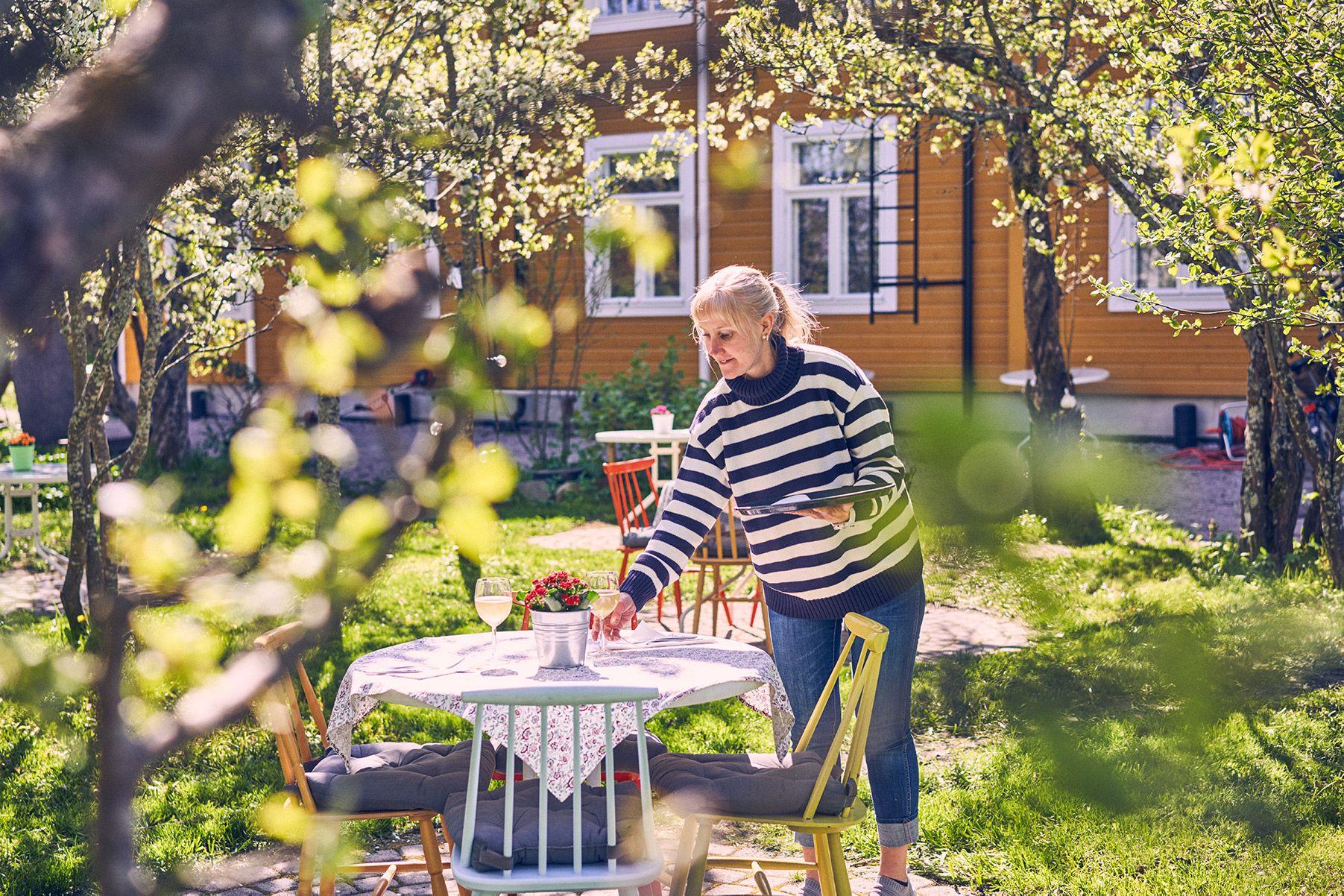 En kvinna med randig tröja dukar ett bord ute. Runt henne finns träd med gröna blad, och i bakgrunden finns ett gult hus.