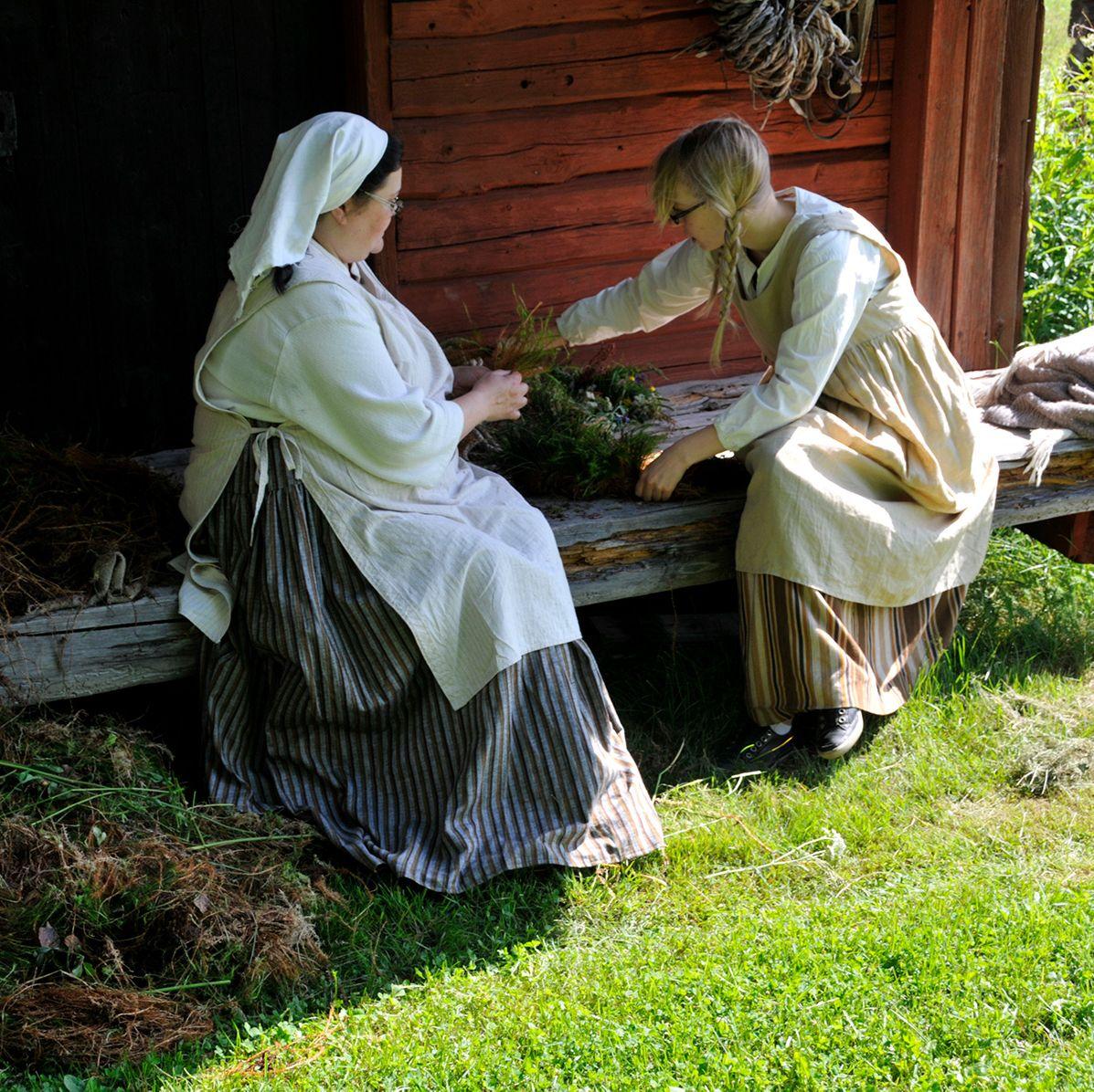 Kaksi naista pukeutuneena vanhanaikaisiin asuihin istuvat punaisen puurakennuksen edessä Sagalundin museon edustalla.